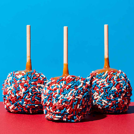 Three candy apples with red, white, and blue sprinkles on a red surface with a blue background