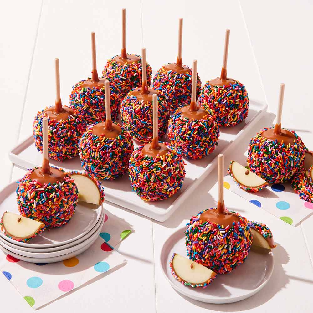 Caramel apples with colorful sprinkles on sticks on a white surface.