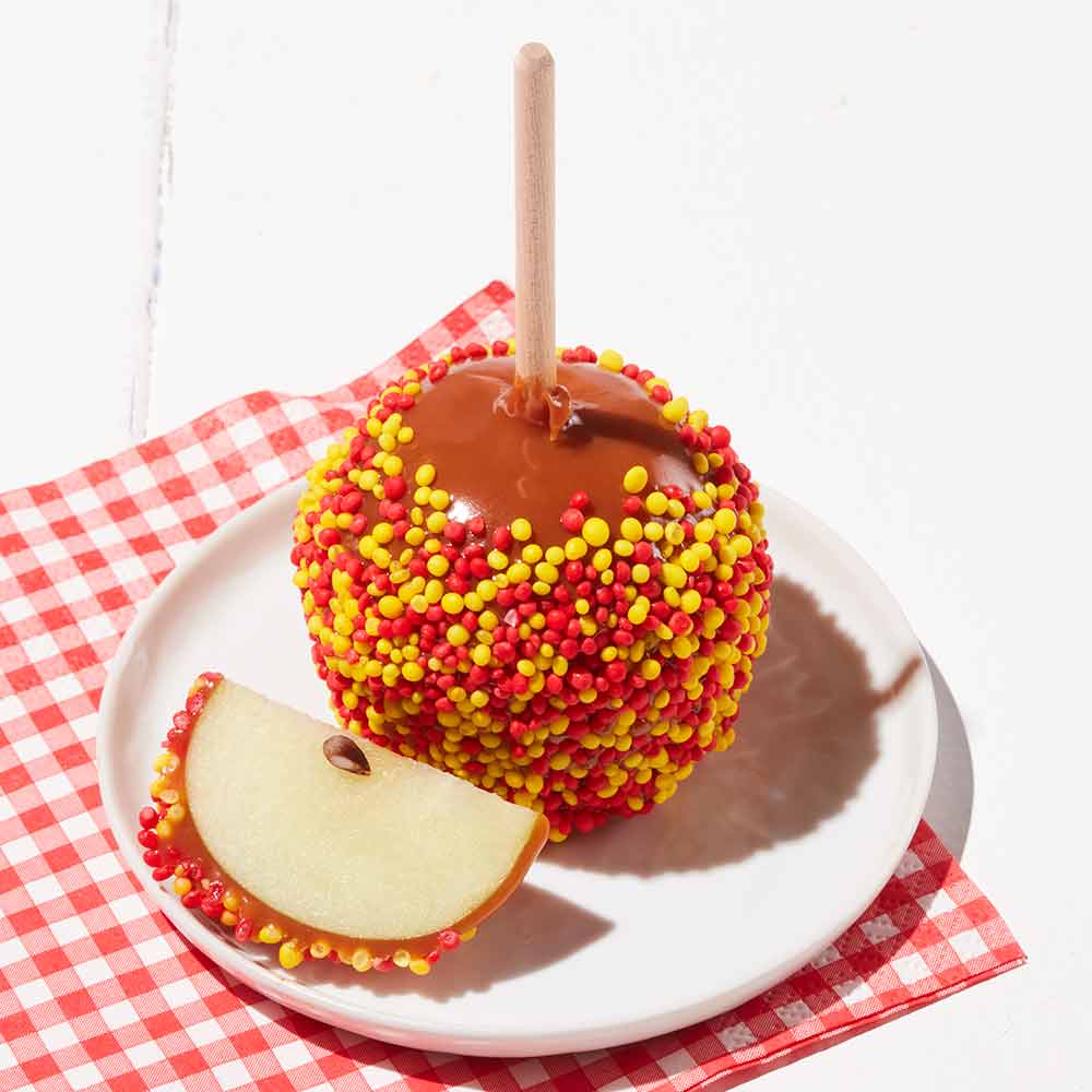 Caramel apple with colorful sprinkles on a white plate with a red checkered cloth underneath.