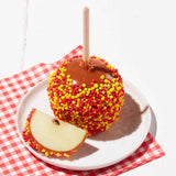 Caramel apple with colorful sprinkles on a white plate with a red checkered cloth underneath.