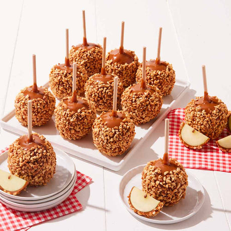Caramel apples with nuts on sticks displayed on a white tray and plates with a red checkered cloth underneath.