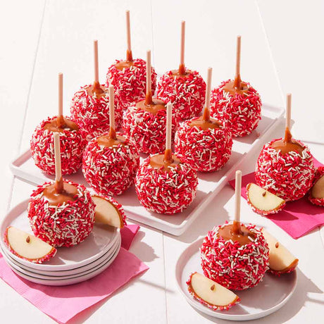 Caramel apples with red candy coating and sprinkles on a white surface.
