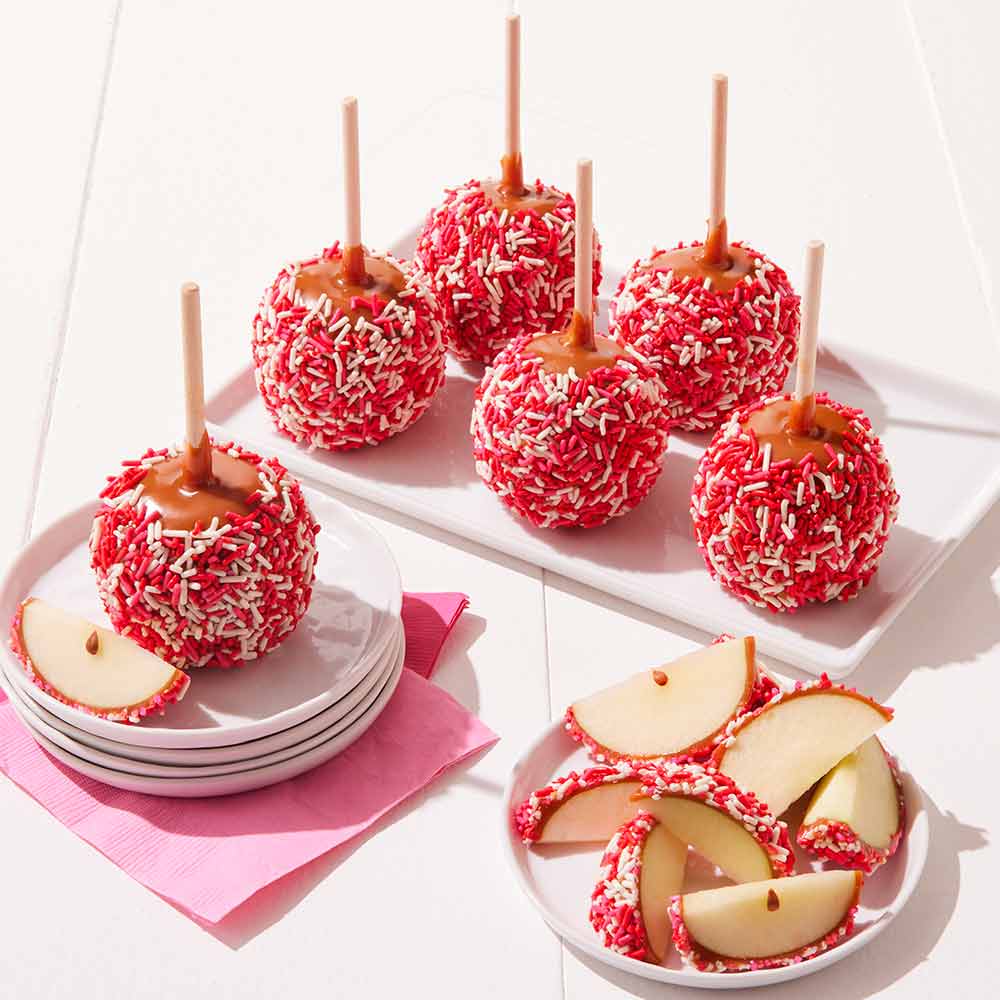 Decorative apples with red and white sprinkles on sticks on a white surface.